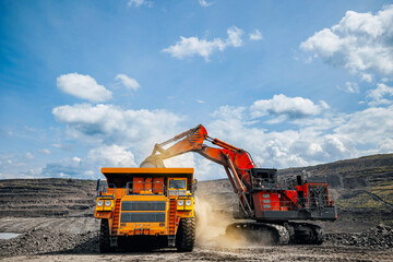 Industrial excavator loads coal into back of yellow heavy dump truck in open pit mine under bright...