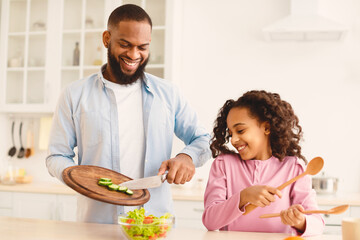 It's Dinner Time. Portrait of black family making healthy meal together to celebrate fathers day, dad adding cucumber slices into glass bowl with knife, girl helping dad and holding wooden utensils