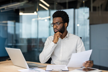 Pensive businessman reading documents and using laptop in office