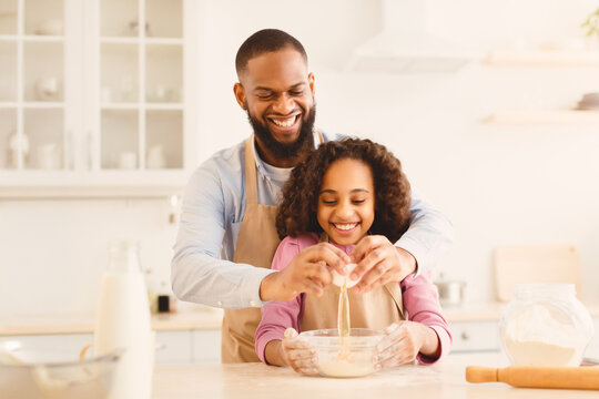 Home Bakery. Excited African American man adding egg to cookies dough, cheerful daughter watching him, learning how to cook together with dad, making pie on father's day, copy space - Powered by Adobe