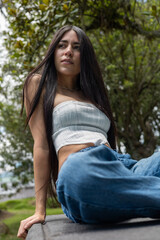 Obraz premium Attractive young woman with long dark hair poses elegantly while reclining slightly backward on a rustic stone bench in a bright garden. Wearing jeans and a white crop top, she conveys confidence, rel