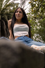 Obraz premium Beautiful young woman with long dark hair sits gracefully on a rustic stone bench in a sunlit garden. Wearing denim jeans and a white crop top, she exudes charm, elegance, and relaxed confidence while