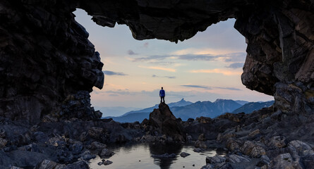 Person Standing Beneath a Rocky Archway Overlooking a Mountainous Landscape