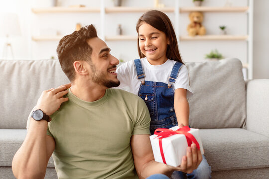 Celebrating Special Occasion. Portrait of smiling cute daughter sitting on sofa at home, embracing dad and greeting him with father's day or birthday, holding and giving wrapped present box