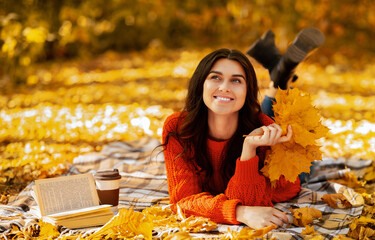 Positive young woman in cozy sweater lying on picnic blanket outdoors with fall leaves, book and takeout coffee. Pretty millennial lady resting on ground in autumn park on right sunny day