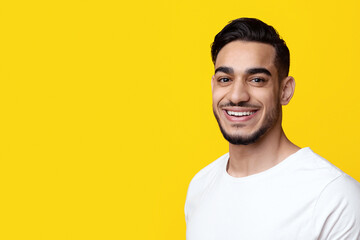 Portrait of cheerful stylish handsome bearded middle eastern young man in white t-shirt posing on yellow studio background, smiling at camera, panorama with copy space. Millennials lifestyle concept
