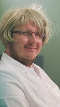 Close-up view of a technician wearing a blonde wig and glasses, smiling at the camera while sitting at a laboratory desk with a microscope
