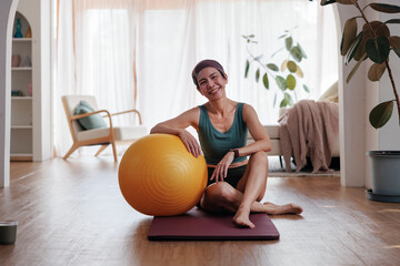 Woman smiles with exercise ball on yoga mat at home.