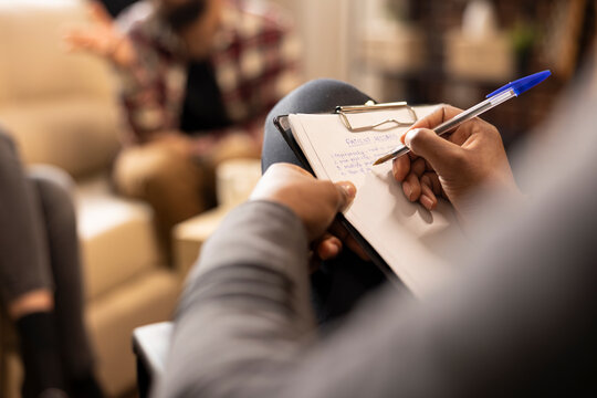 Close up of African American hands writing with pen on paper, documenting session in office. Black male therapist seated with clipboard, attentively taking notes during consultation.