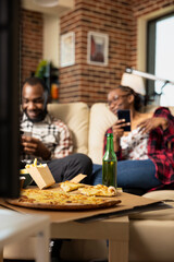 Closeup of pizza on table as boyfriend and girlfriend enjoy cozy time at home. African american couple digital bonding on weekend with takeaway food nearby in brick wall apartment.