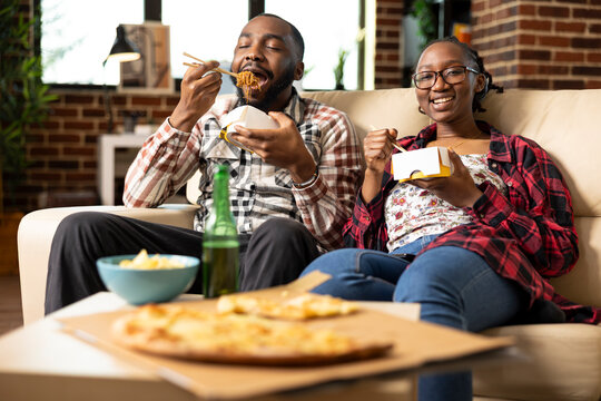 Young black couple relaxes at home, watching television together with takeout food. African american boyfriend eating noodles with girlfriend, enjoying downtime in brick wall apartment. - Powered by Adobe