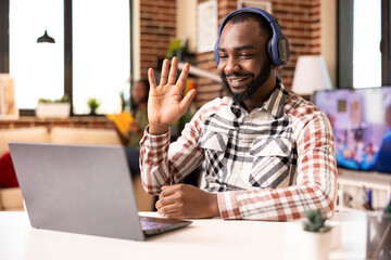African American male freelancer gestures hello on video call, attending remote skills workshop. Friendly man with headphones greets remote colleagues during virtual training session.