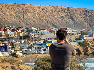 Back view of a male tourist taking photos of charming coastal town of Imsouane. Young man stands on...