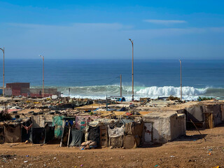 Coastal settlement in Imsouane with small shacks constructed from various materials and surrounded by junk and debris. Beach and ocean with beautifully breaking waves are visible in the background.