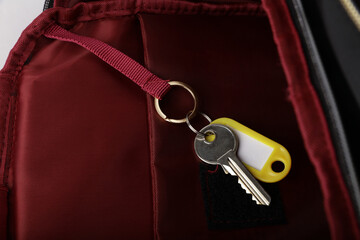 A close-up of a key and a key tag hanging from a red ribbon loop inside a dark-colored backpack. The image highlights a smart and simple way to organize and easily locate keys.