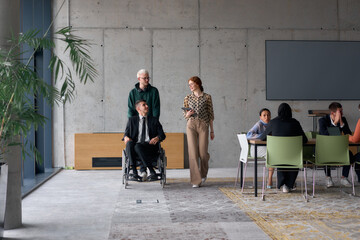 A diverse team of business colleagues, including a director in a wheelchair, engages in a collaborative discussion as they move together through a modern office space