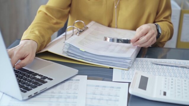 Female auditor in business attire inspecting financial reports with magnifying glass, surrounded by documents and a laptop and calculator. Business audit and taxes concept