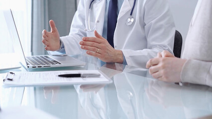 Doctor is consulting patient in modern fair clinic. Healthcare professional discusses medical information with a woman, gesturing above the laptop computer on the glass desk. Medicine
