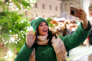 Excited European woman making selfie or having video call outdoors while walking on Xmas market and...