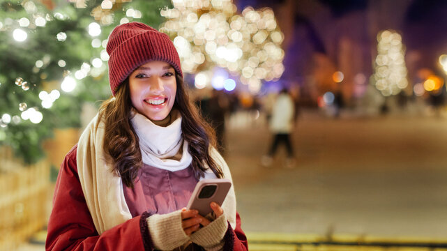 Lady using cellphone on Christmas market while walking and enjoying festive winter season and vibe, blurred decorated fir tree with lights on background