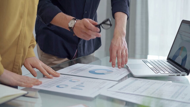 Business people, professionals analyzing financial charts and data on a desk while standing and using laptop computer in office. Dark blue shirt and yellow blouse