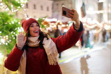 Excited European woman making selfie or having video call outdoors while walking on Xmas market and showing festive Christmas fair atmosphere