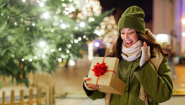 Excited surprised lady opening gift box and rejoicing, standing outdoors in front of decorated Xmas tree with garland lights, free space - Powered by Adobe