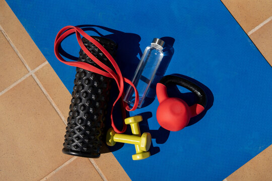 Set of fitness equipment on yoga mat including dumbbells, kettlebell, resistance band, foam roller, and water bottle outdoors
