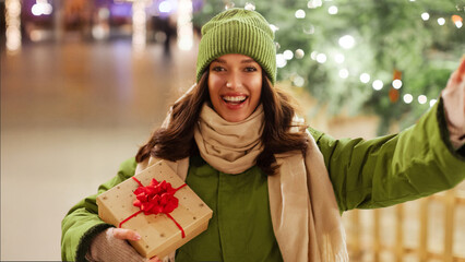 Happy young European woman with gift boxes in hands posing outside on Christmas Eve, taking selfie...