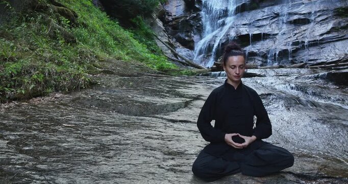 Aerial drone shot of woman practicing slow tai chi movements or meditating yoga near a waterfall. Spreading arms, expressing harmony, sitting in lotus position, mindfulness, fitness and spiritual