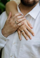 Close up of couple's hands with wedding and engagement rings on white shirt background
