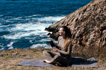 Woman practicing meditation and stretching on yoga mat by the ocean, surrounded by rocky cliffs and waves, embodying tranquility and mindfulness in nature