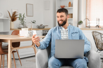 Young bearded man working with laptop and mobile phone in armchair at home