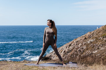 Woman in form-fitting activewear practicing stretching and meditation on a yoga mat by the ocean, surrounded by natural beauty and tranquility, promoting wellness and mindfulness