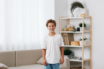 Smiling boy stands indoors in a bright, minimalist living room with white shirt and jeans, creating...