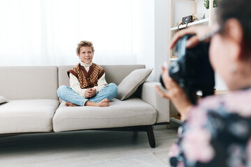 Obraz premium Authentic play scene featuring a young boy sitting cross legged on a sofa, dressed in playful school style, as a photographer captures candid moments at home.