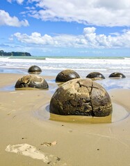 Coastal scene, large, dark-brown spherical rocks on sandy beach, water lapping, blue sky