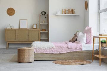 Bed with stylish pink bedding, wicker table, commode, shelving unit and houseplants in bedroom at home