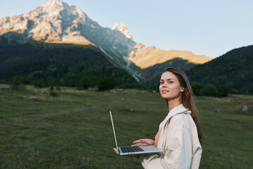 A young woman outdoors holds a laptop while standing in a grassy field with distant mountains. She appears focused and confident, creating a calm, productive work moment.