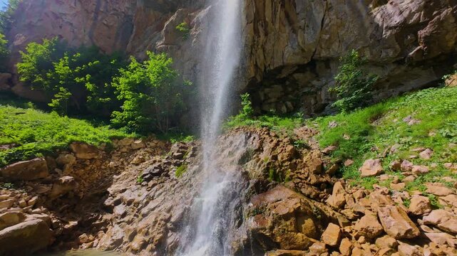 Aerial video of a scenic waterfall cascading down a rocky mountain cliff surrounded by lush green trees and forest, captured in smooth FPV drone slow motion on a sunny summer day.