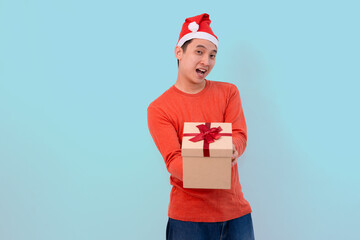 A young man wearing a red Santa hat joyfully offers a wrapped gift box in christmas concept blue background
