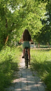 Back view of young woman confidently riding bicycle through a lush green park path, her long hair flows freely, blending with natural scenery, background includes trees and a building
