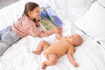 Cute little girl reading story to her sleeping baby brother on bed, top view