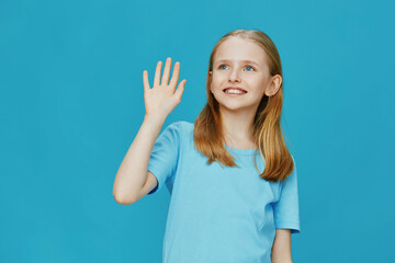 Happy young girl waving on a bright blue background, wearing a light blue shirt, showcasing joy and...
