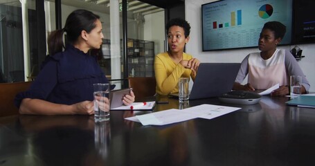 Three businesswomen examining charts and data in conference room with laptop tablet speakerphone