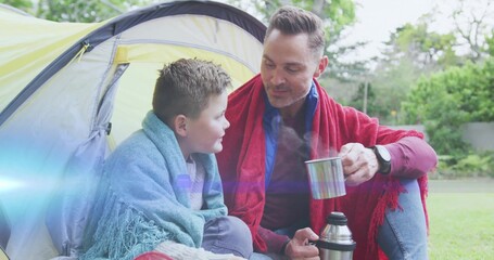 Holding mugs wrapped in blankets, father and son chatting at green campsite with tent, thermos