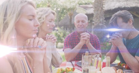 Praying family bowing heads and clasping hands around checkered table in garden, with skewers