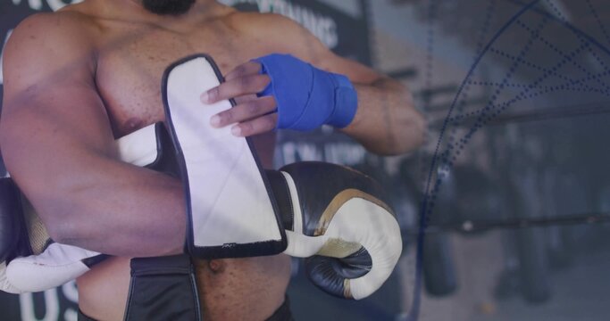 Wrapping shirtless boxer applying blue hand wraps holding white gloves at boxing gym, punching bags - Powered by Adobe