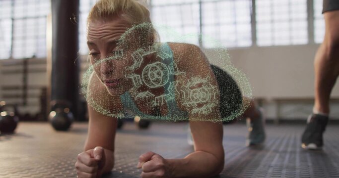 Woman wearing sportswear holding forearm plank on mat in gym, with hologram gears and kettlebells - Powered by Adobe