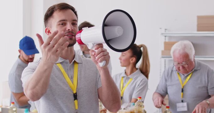 Young man volunteer screaming in megaphone working at charity center announcing free food donations of foodstuffs for poor people in charitable foundation. Humanitarian aid concept. 4k video.